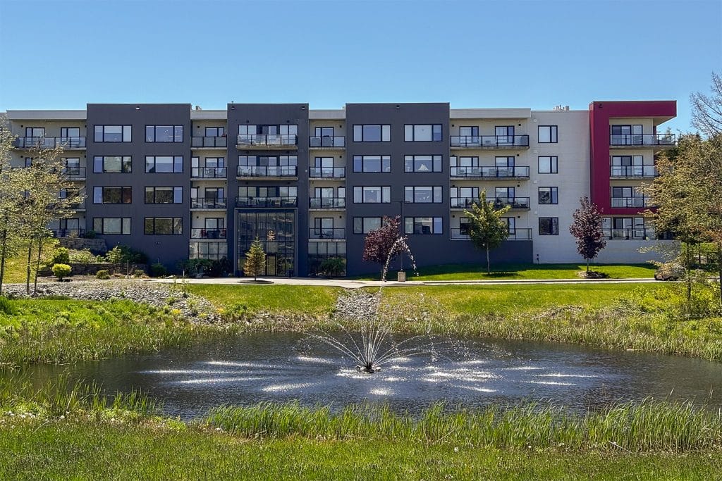 Shiraz Gardens Amarone apartment building with a pond with a fountain surrounded by trees and sunny grass. This building is located in Hammonds Plains, Nova Scotia